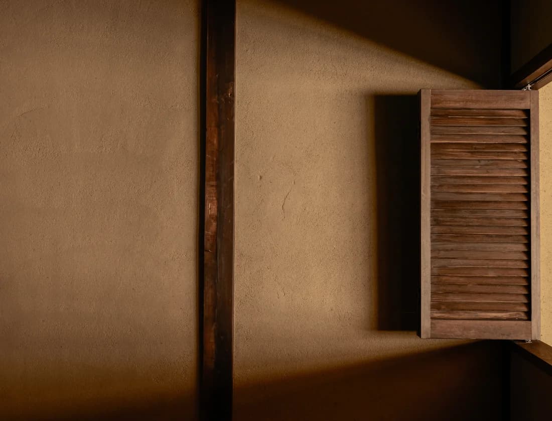 Wooden louvered shutters filtering light against a clay plaster wall